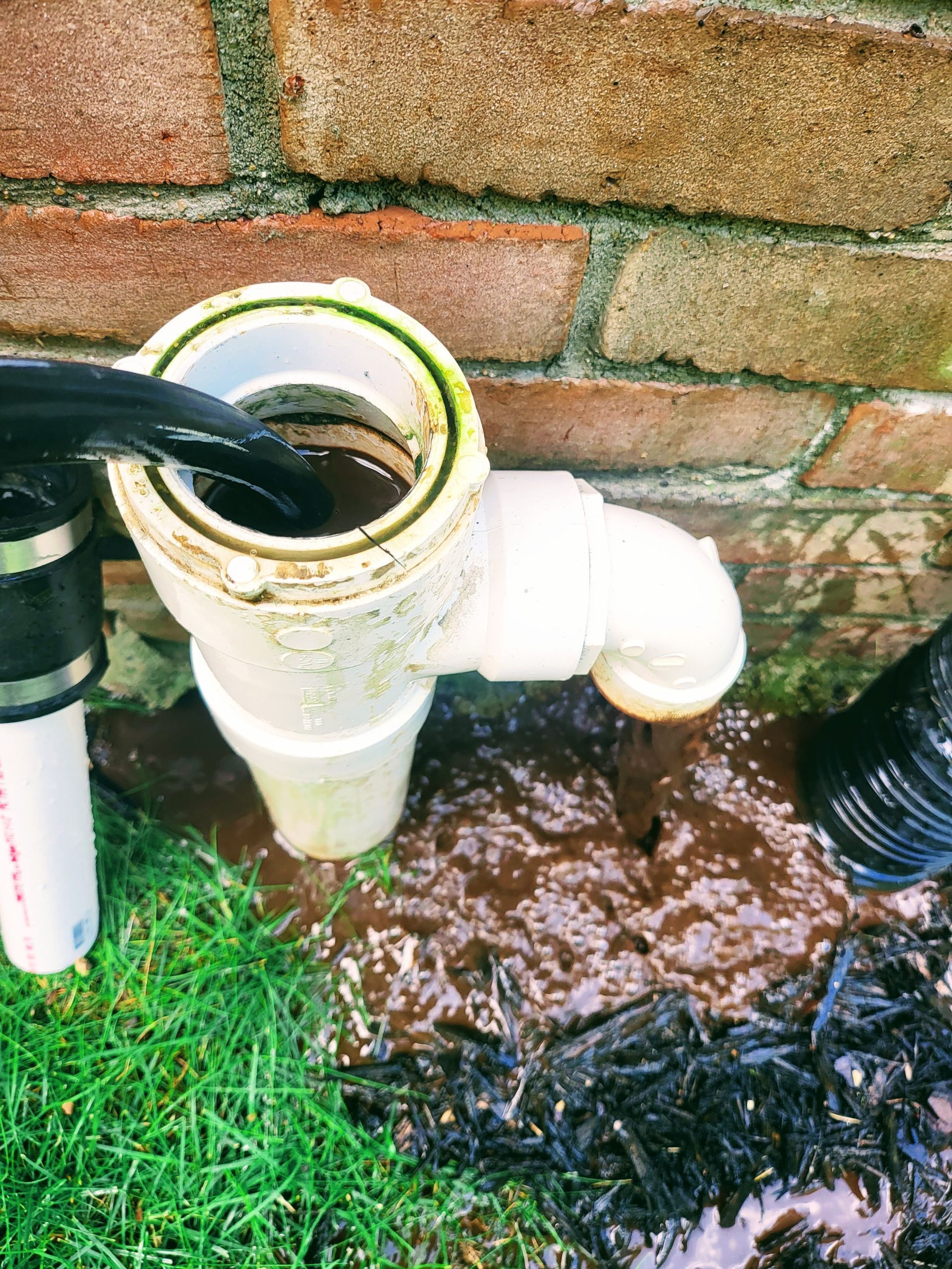 A pipe with water coming out of it is sitting in the grass next to a brick wall.
