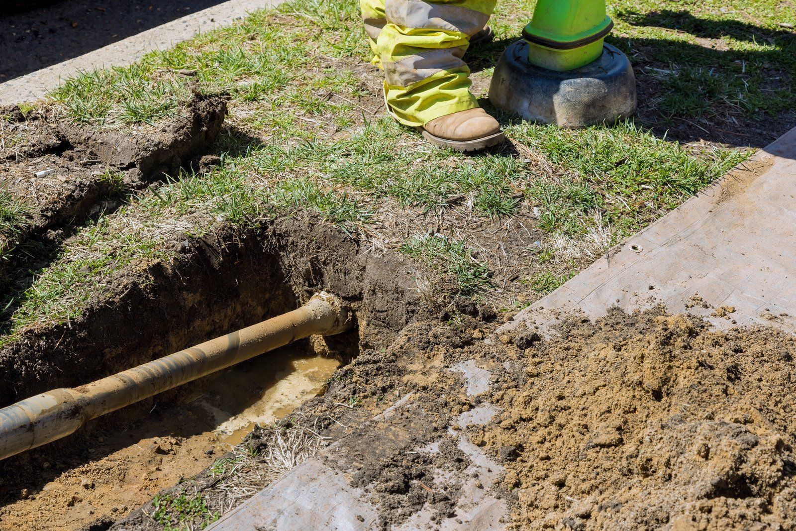 A worker in yellow boots excavates near a concrete sidewalk, digging around a pipe in a grassy area.