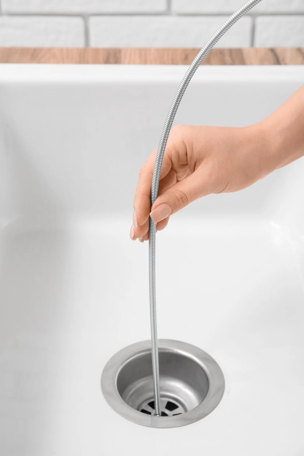 A hand inserts a flexible, metal drain snake into a white kitchen sink drain to clear a blockage.