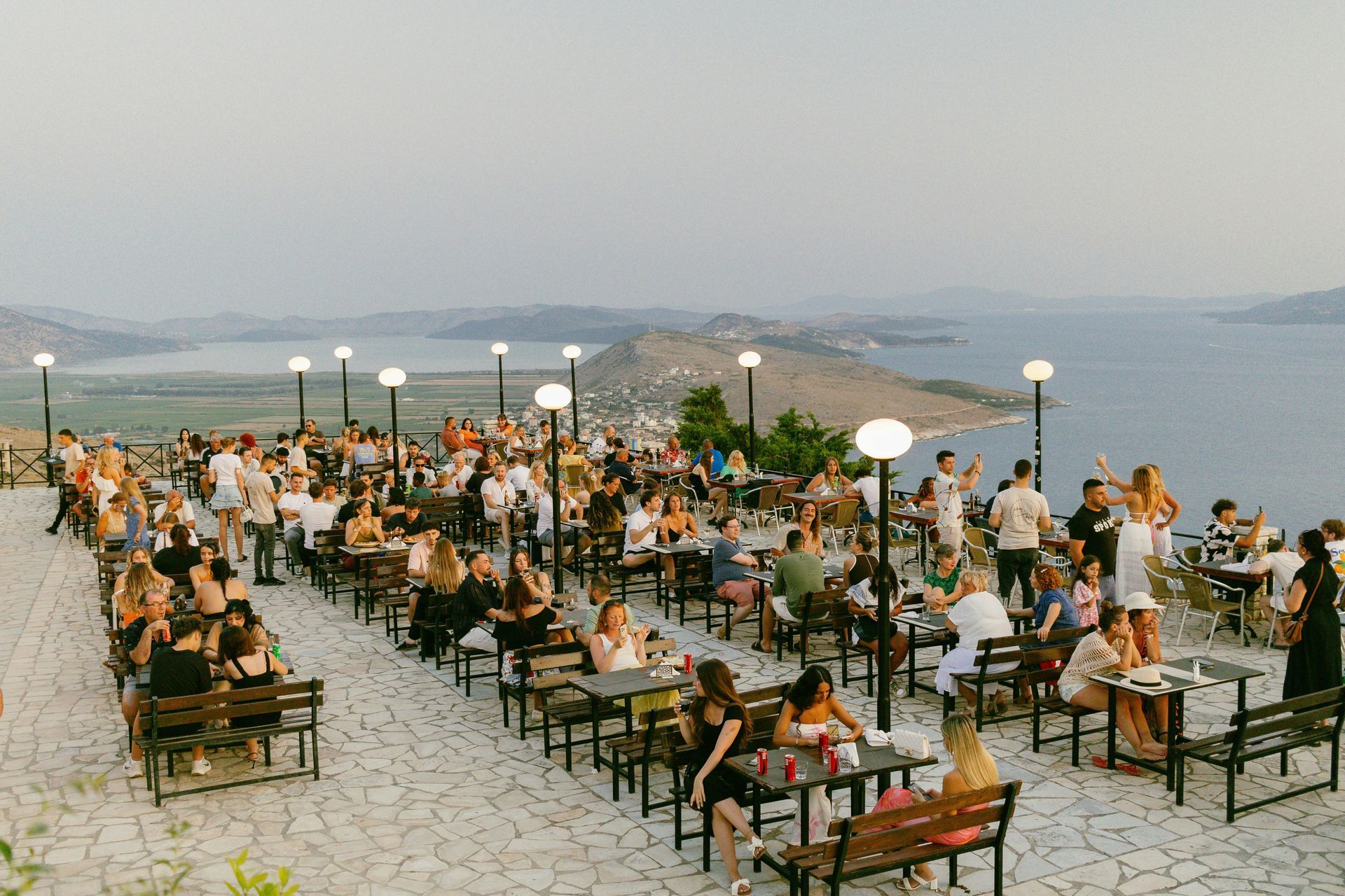 People on a terrace overlooking the sea at sunset. Some are seated, enjoying drinks in Saranda