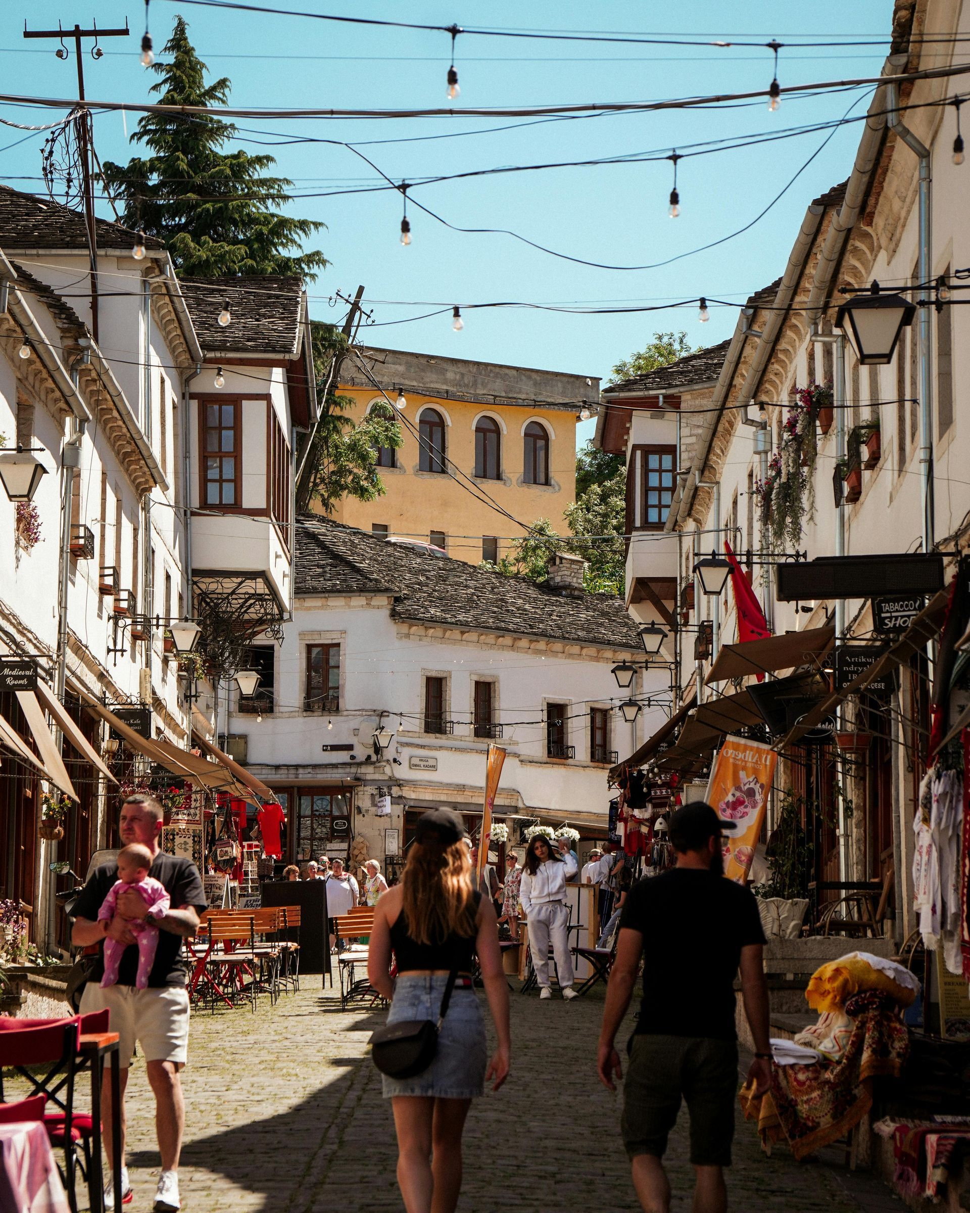Narrow street in a European town with shops and people walking. Buildings are white with brown roofs. Bright blue sky.