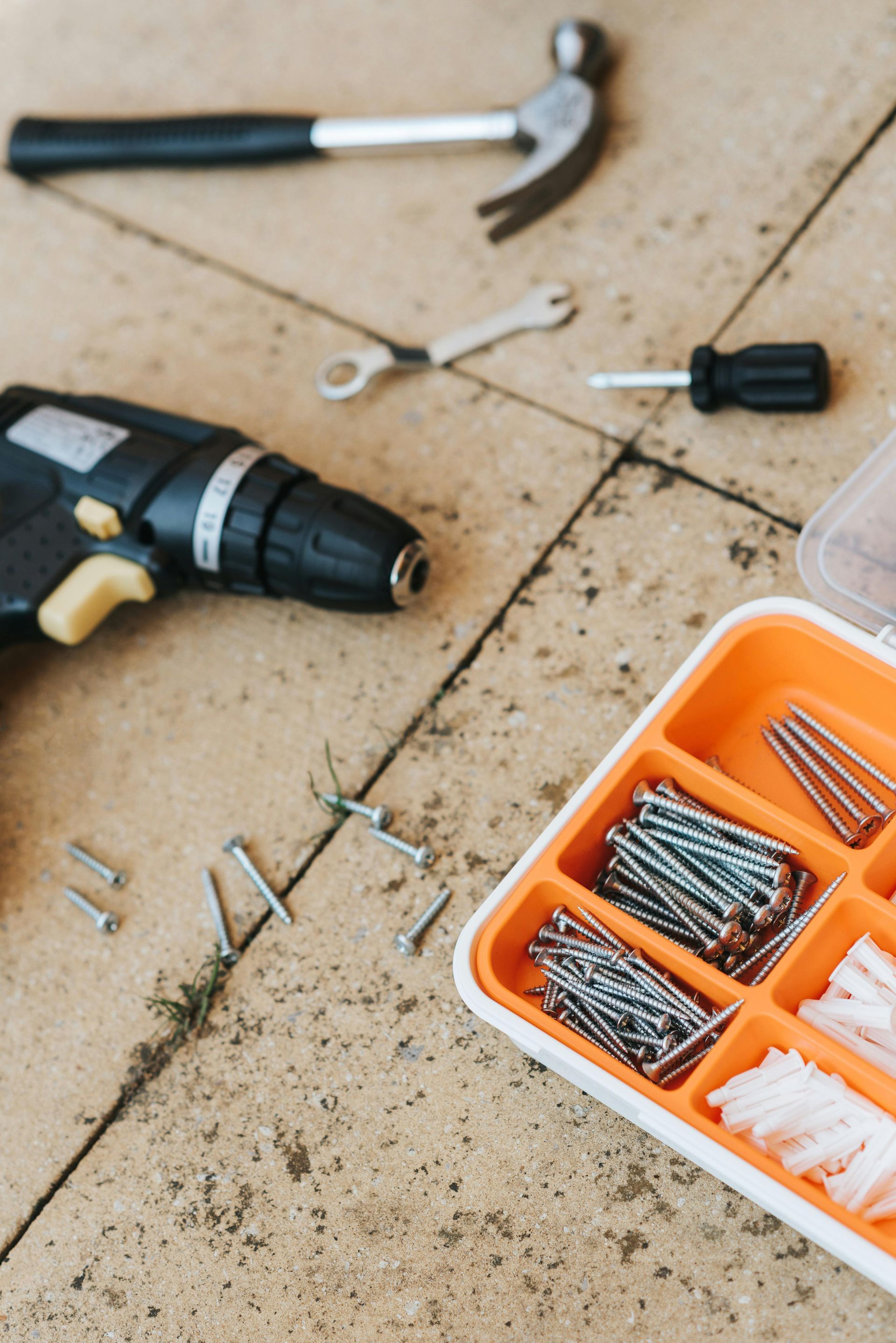 Tools on a tiled surface, including a drill, hammer, screwdriver, wrench, and screws in a divided container.