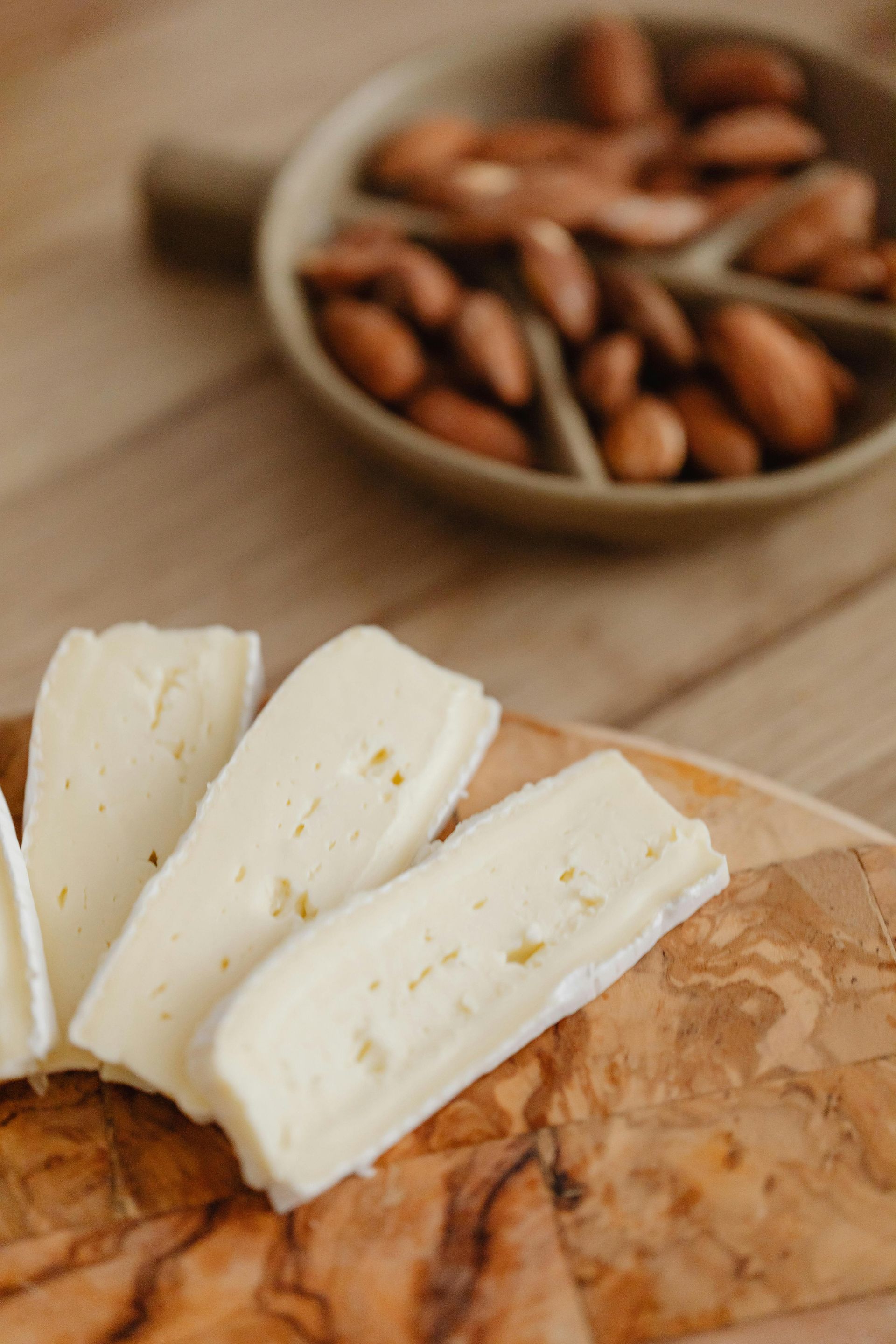 Sliced brie cheese on a wooden board, with a dish of almonds in the background.