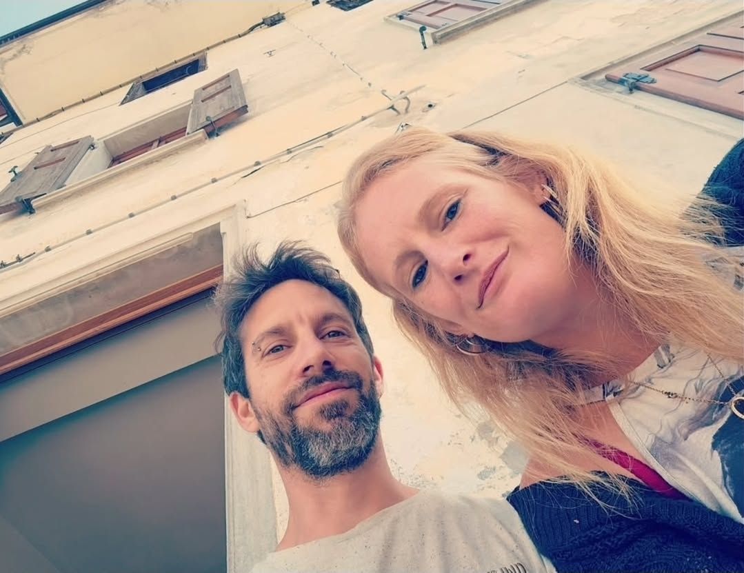 Een lachend stel poseert voor een selfie buiten een wit gebouw met rode luiken.