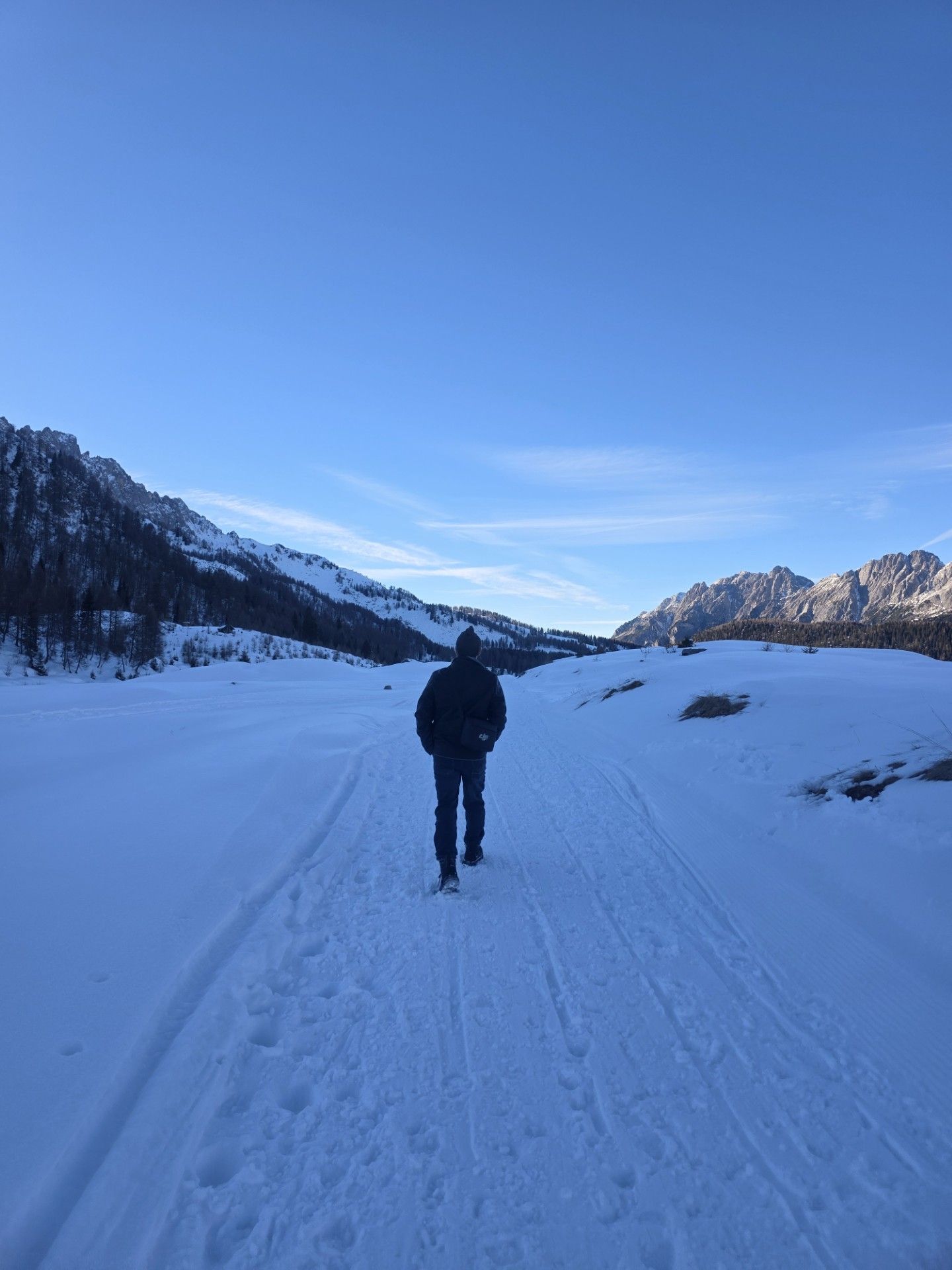 Een persoon loopt over een besneeuwd pad richting de bergen onder een blauwe hemel.