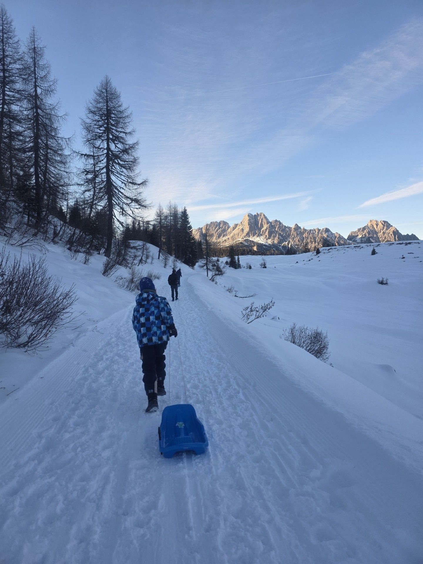 Kind trekt een blauwe slee over een besneeuwd pad; bergen en bomen op de achtergrond.