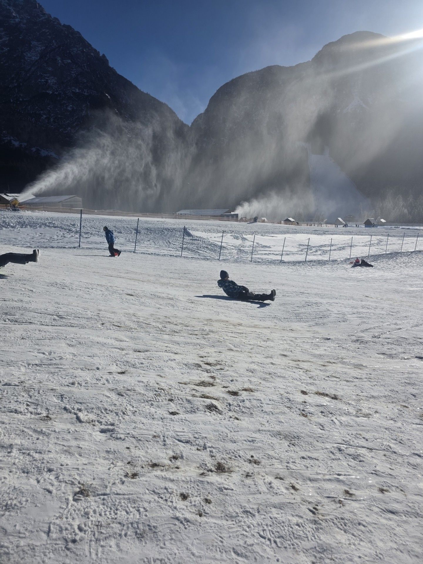 Besneeuwd landschap met sneeuwkanonnen die sproeien, een persoon die in de sneeuw ligt. Bergen op de achtergrond, zonnig weer.