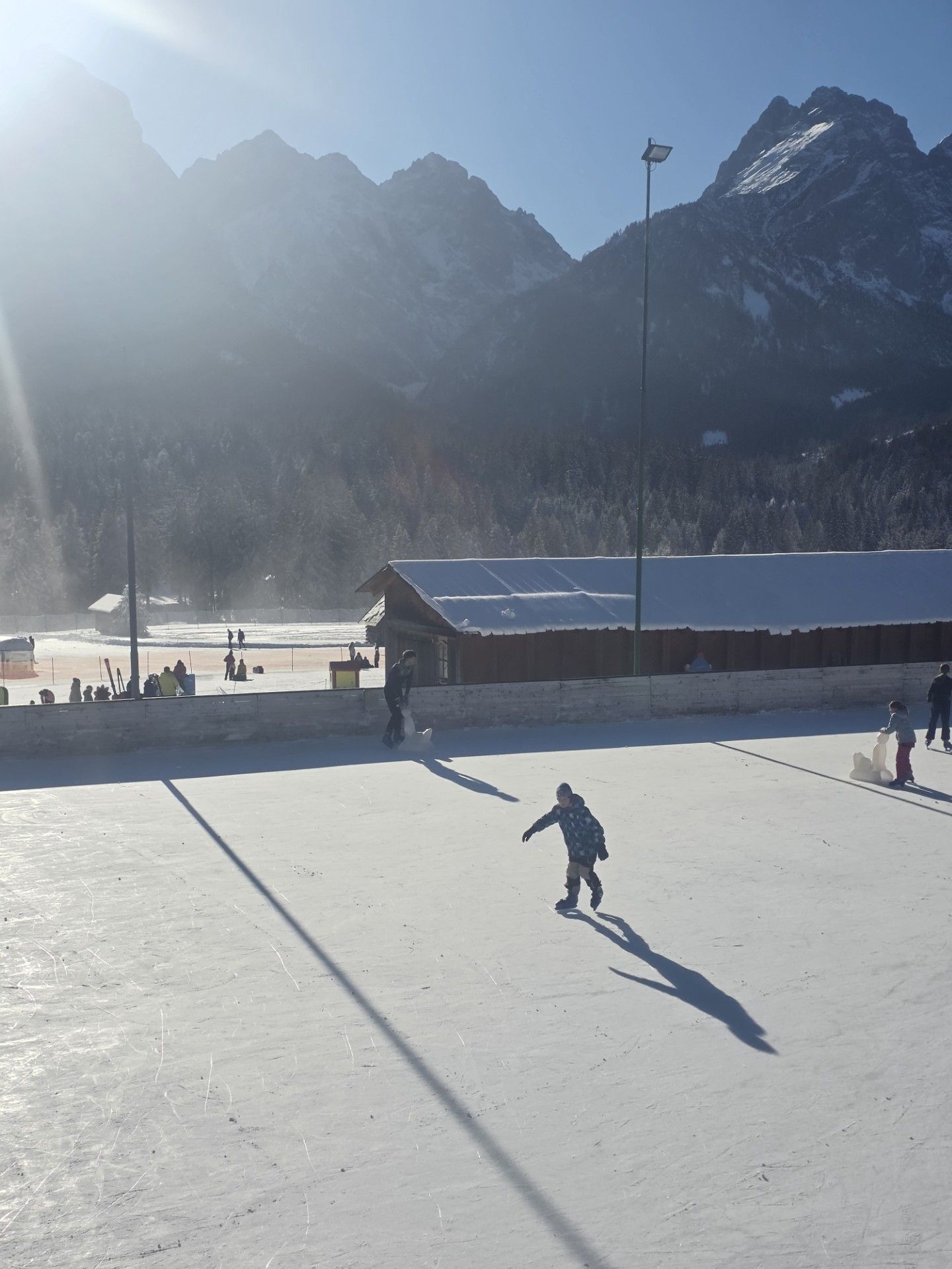 Een persoon schaatst op een ijsbaan met bergen op de achtergrond, in een stralende zon.