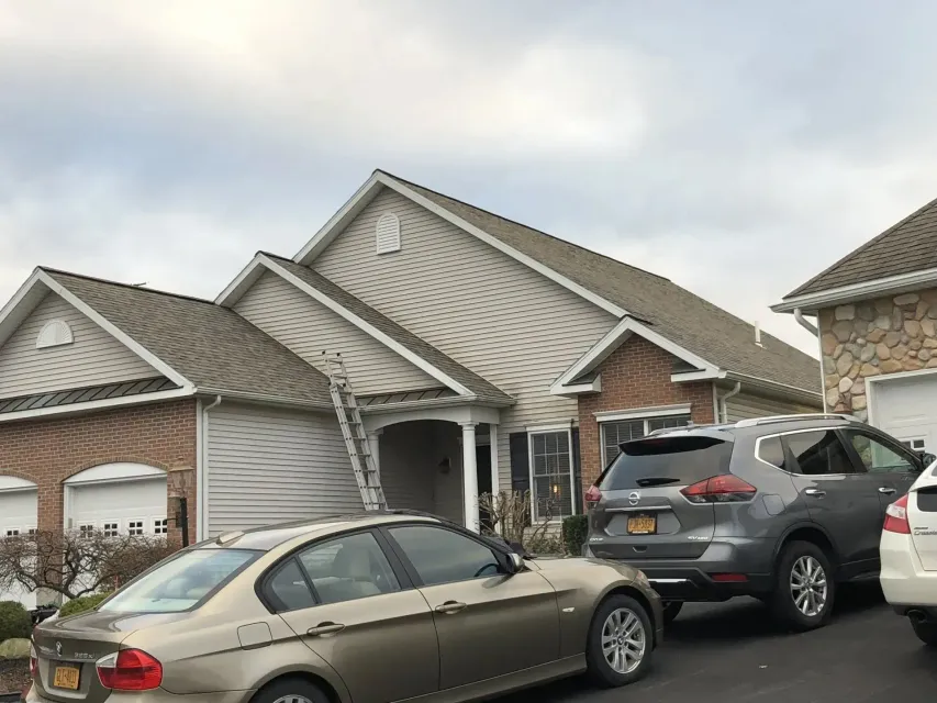 Beige house with gray siding, a brick facade, and parked cars in the driveway. A ladder leans against the house.