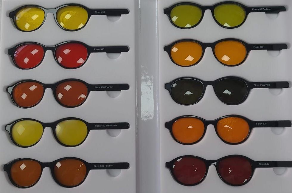 Un paio di occhiali geometrici esposti su uno scaffale rosa in un negozio. Montatura nera con lenti trasparenti e un'immagine promozionale sullo sfondo.