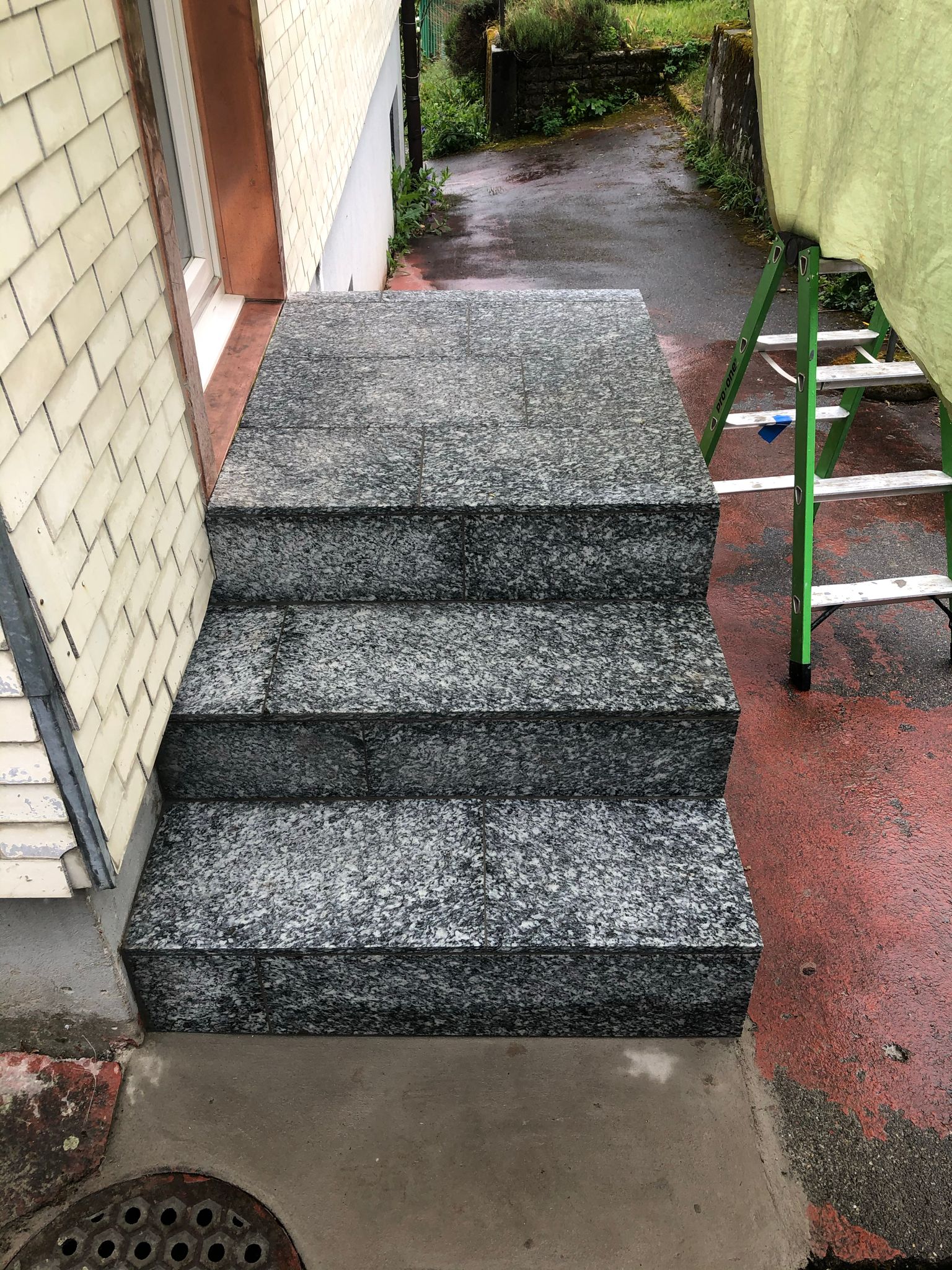 Eine Treppe führt zu einem Haus hinauf. Im Hintergrund ist eine grüne Leiter zu sehen.