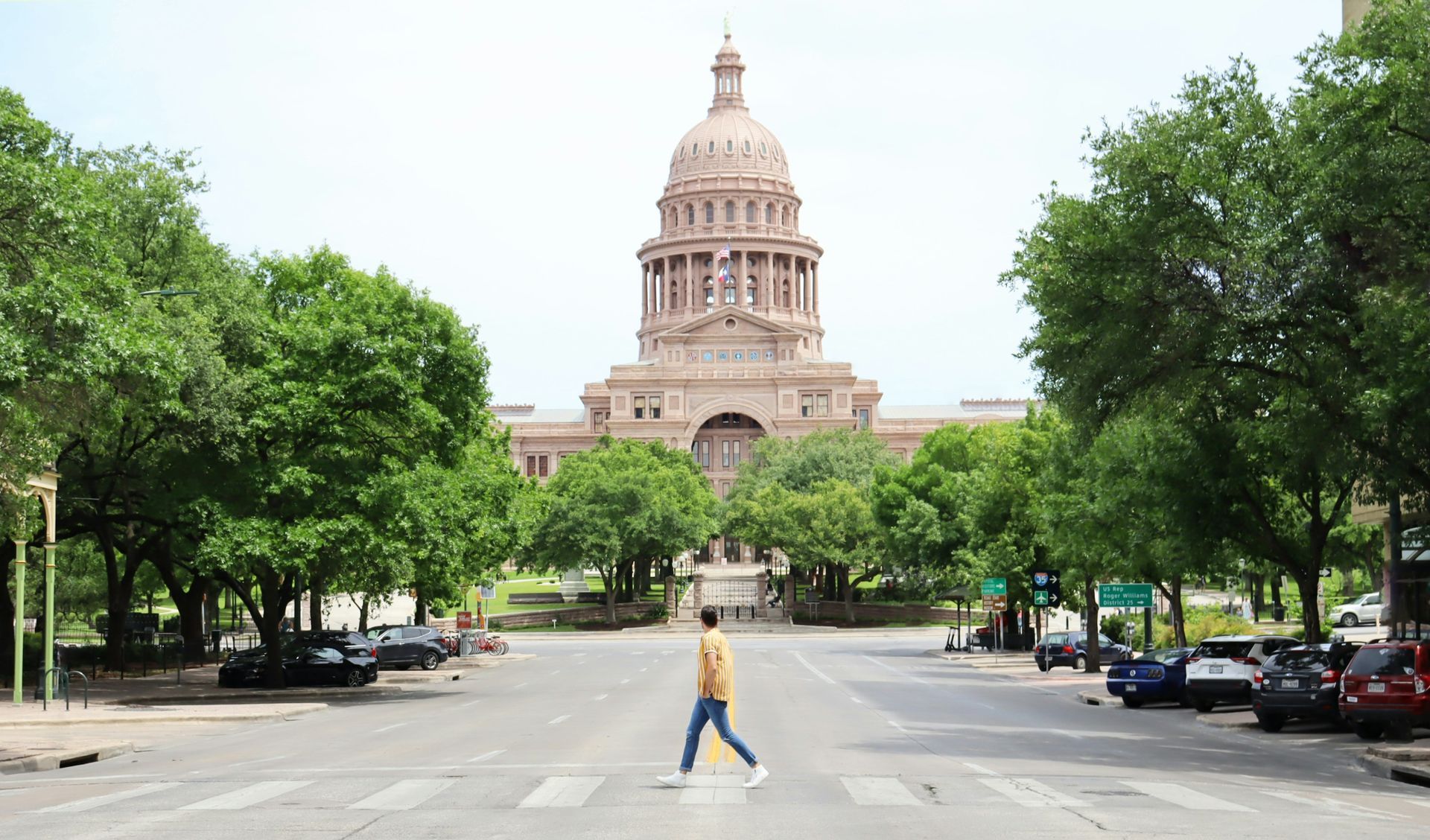 Person walking across a street towards the Texas State Capitol building, with cars parked on either side, sunny day.