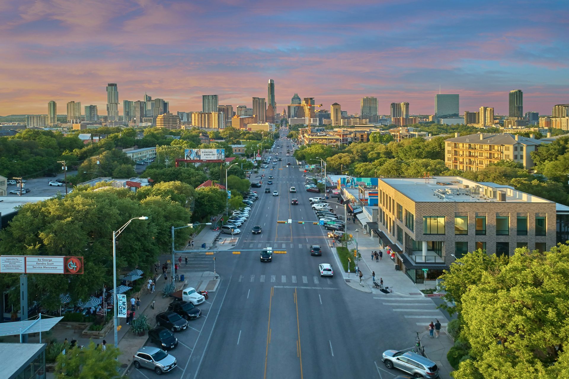 City street with cars, buildings, and trees under a colorful sunset sky; Austin, Texas.