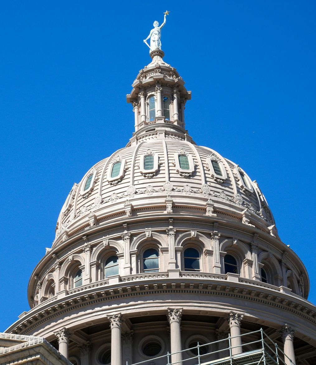 Texas State Capitol dome, tan stone with windows, topped with a statue against blue sky.