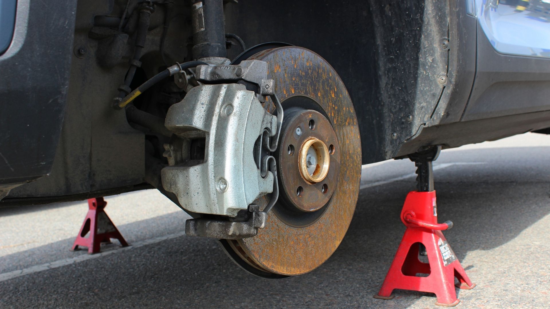 Car raised on jack stands; wheel removed, exposing brake rotor and caliper. Rusty metal and red stands.