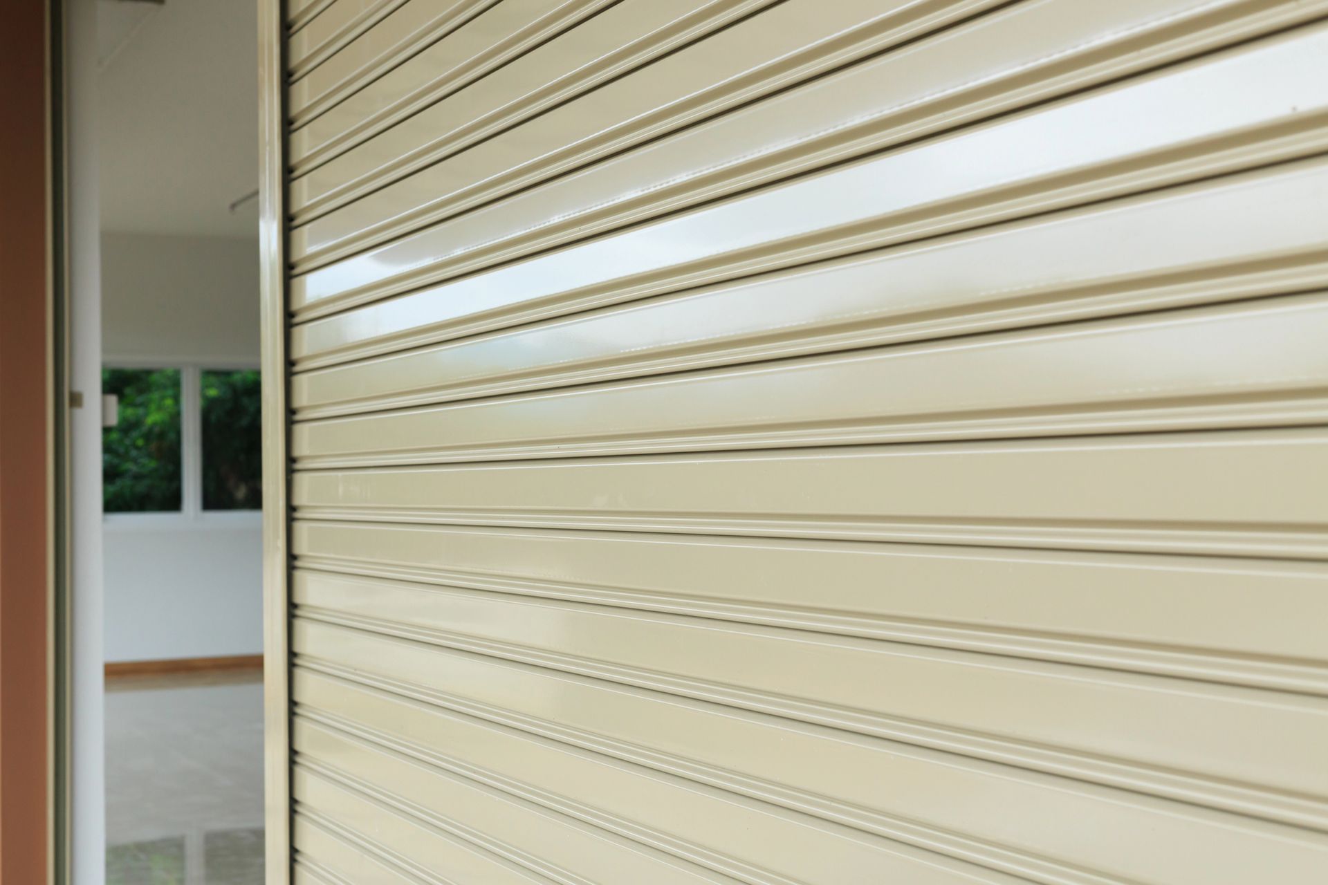 Porta avvolgibile beige parzialmente aperta, che rivela una stanza piena di luce con una finestra e pareti bianche.