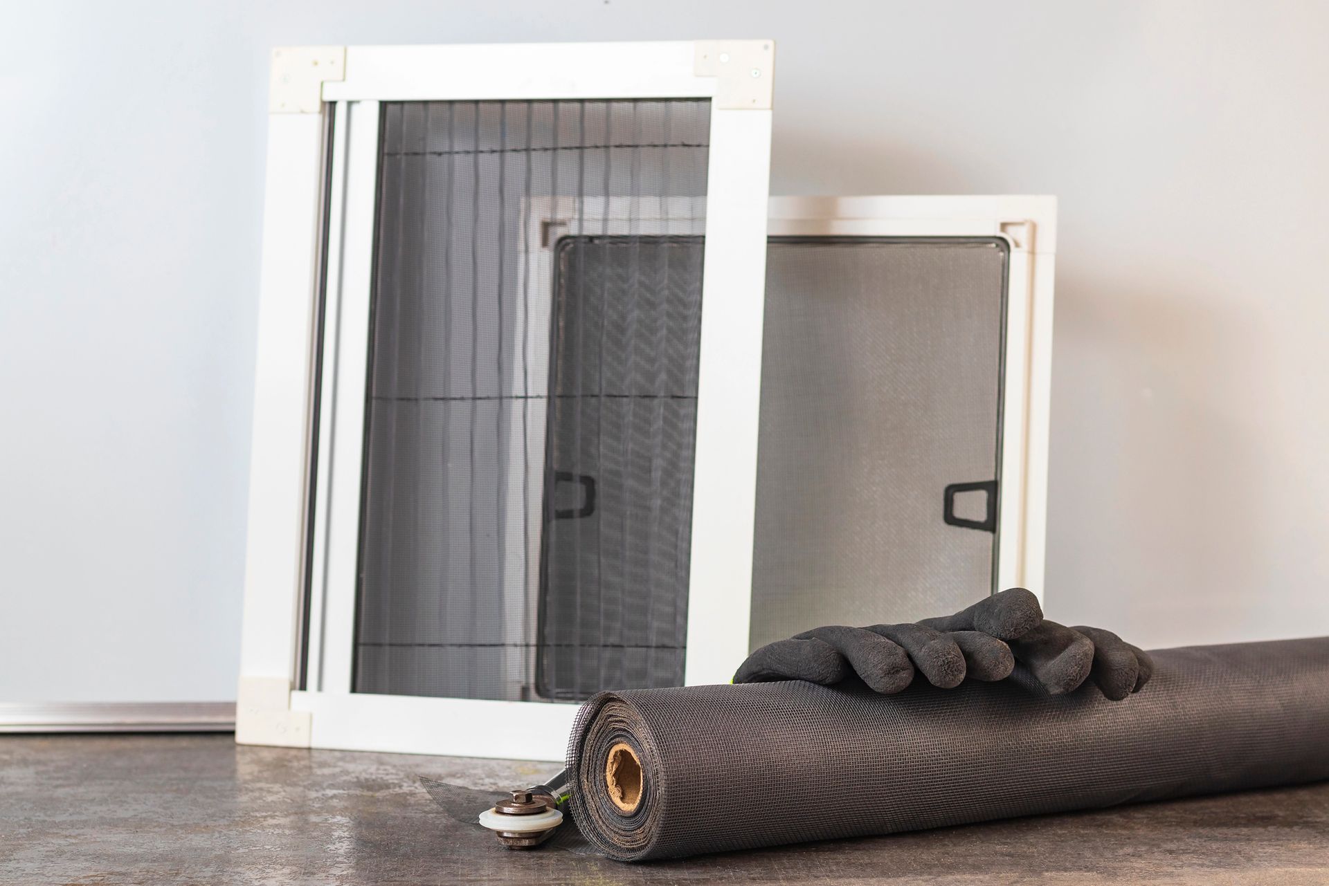 Due zanzariere con cornice bianca, un rotolo di materiale per zanzariere e guanti su una superficie, probabilmente per la riparazione della zanzariere.