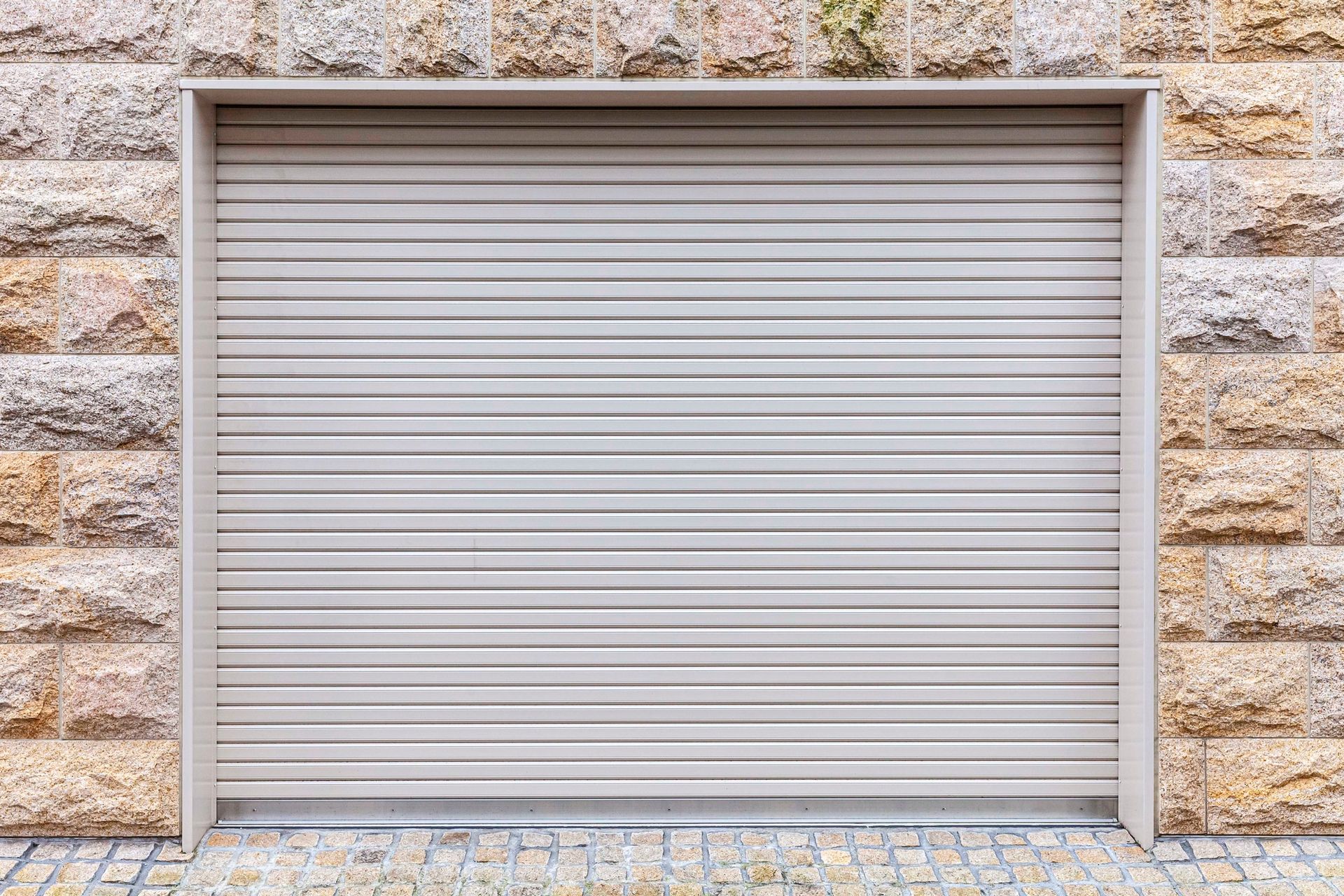 Porta da garage avvolgibile beige chiusa, inserita in un muro di pietra color marrone chiaro.