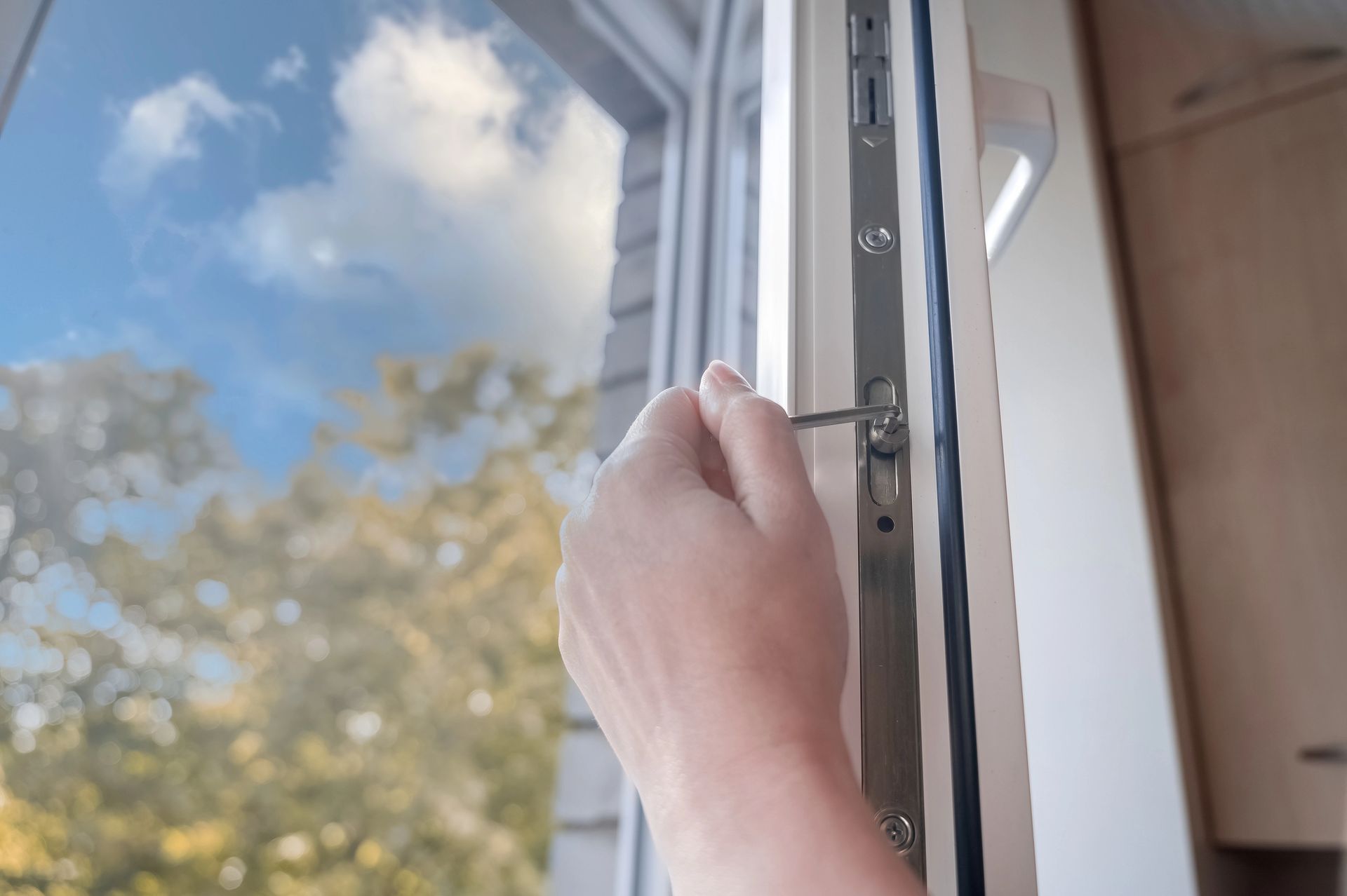 Mano che usa uno strumento per regolare il meccanismo di chiusura di una finestra. Cielo azzurro e alberi visibili all'esterno.