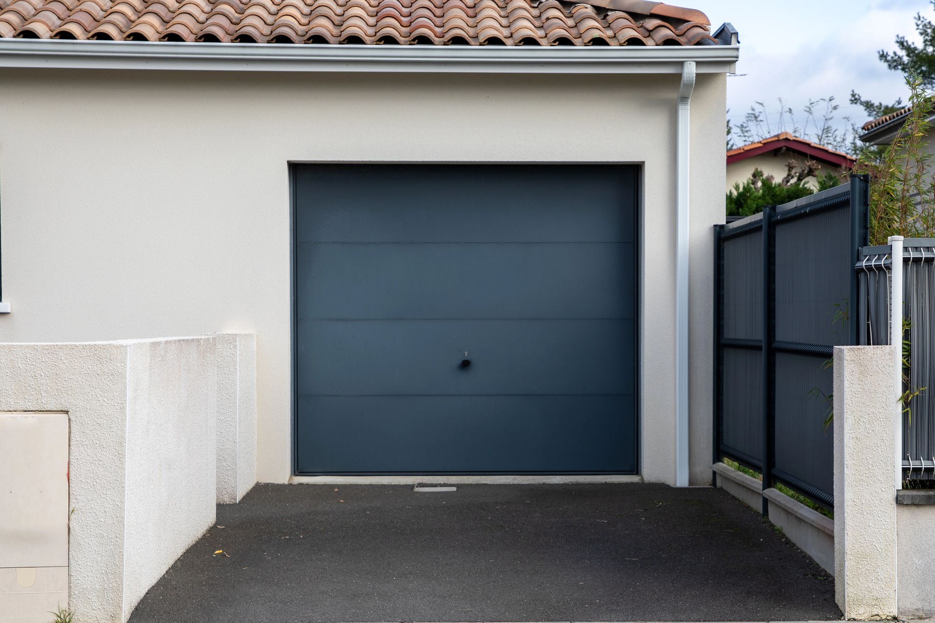 Porta di garage grigia su un edificio di colore chiaro, situata su un vialetto d'asfalto scuro; adiacente recinzione scura.