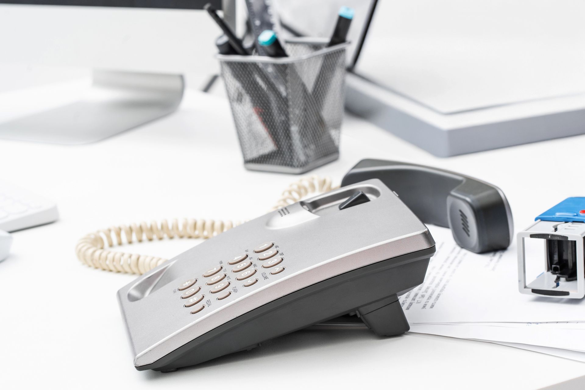 Telefono fisso argentato su una scrivania bianca, con penne e un documento nelle vicinanze.