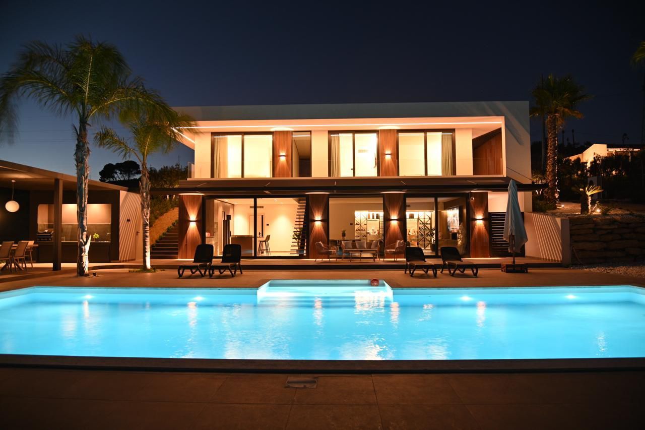 Casa moderna con finestre illuminate di notte, con vista su una piscina blu.