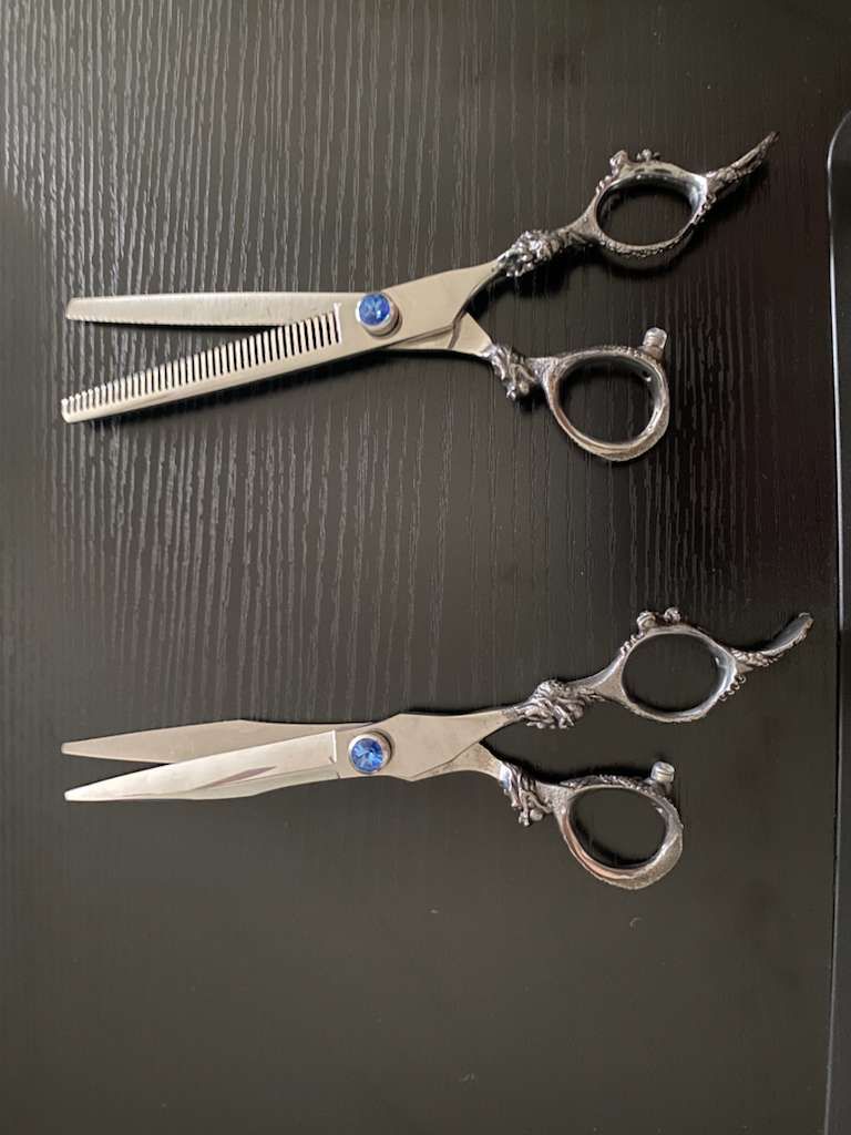 Two ornate silver and blue hairdressing shears on a dark surface. One is a thinning shear.
