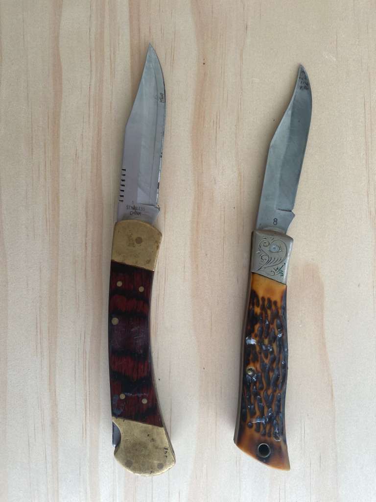 Two pocket knives on a light wood surface; one with a wood and brass handle, the other with a brown textured handle.