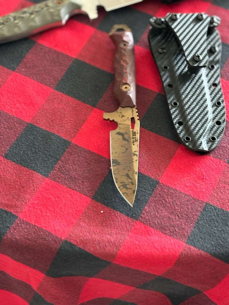 Knife with brown handle and textured blade on red and black checkered cloth, next to a black sheath.