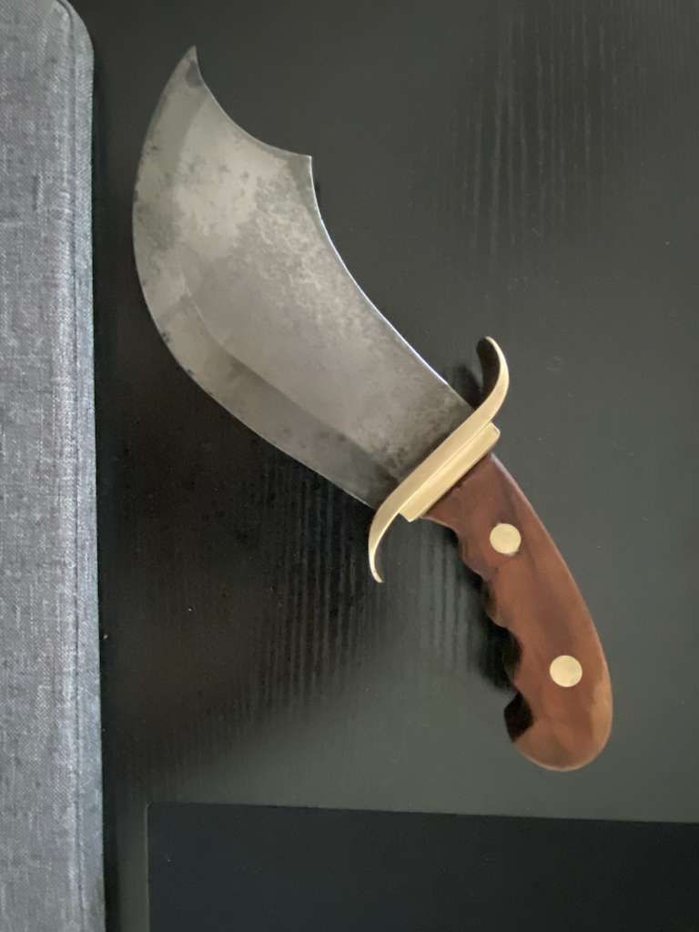 Large, curved blade knife with wooden handle and brass guard mounted on a black surface.