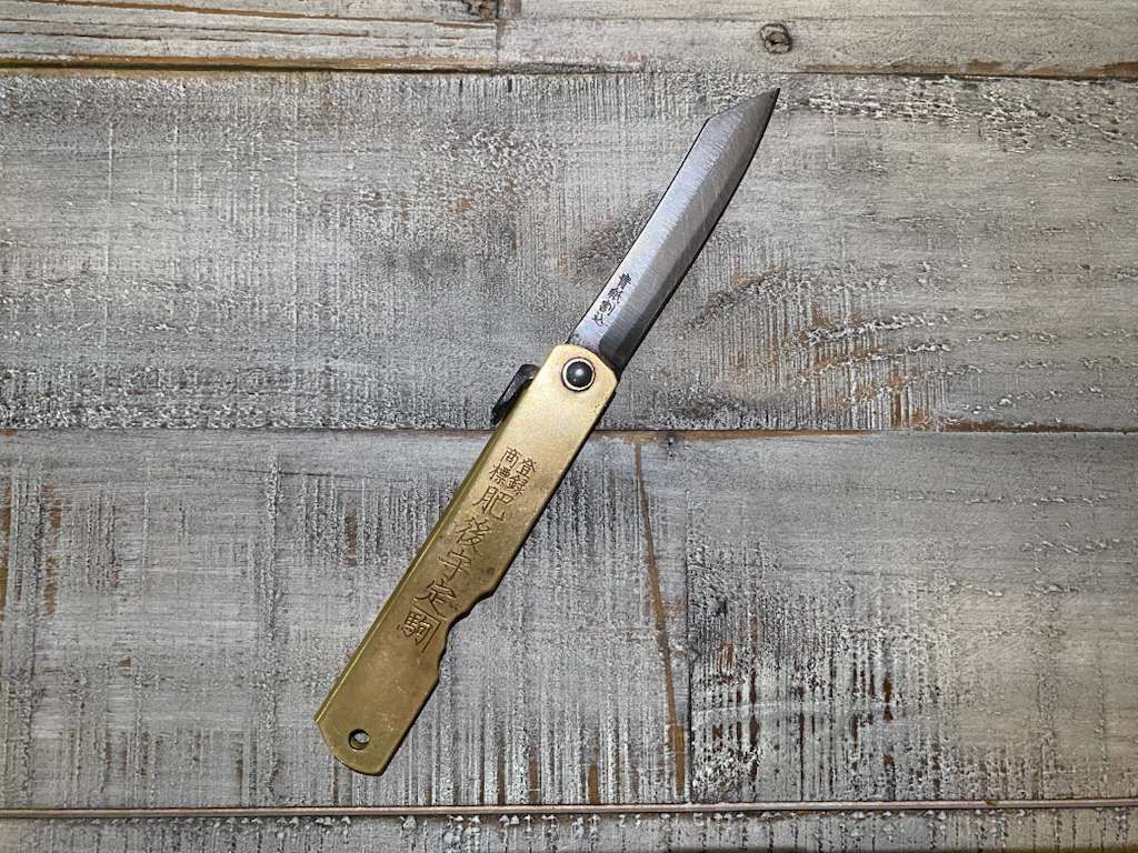 A closed folding knife with a brass handle and a partially serrated blade, on a weathered wooden surface.