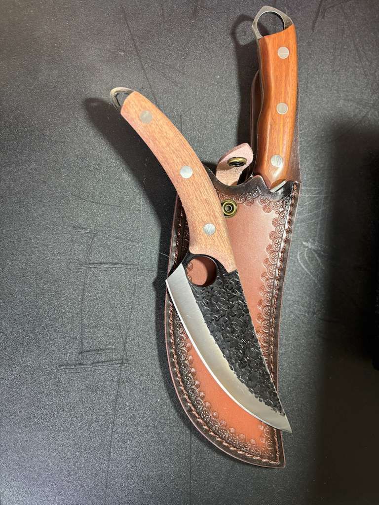 Two knives with wood handles and leather sheath on dark surface.