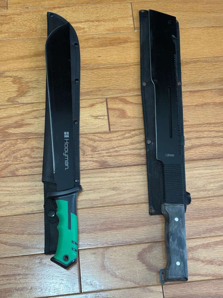 Two black machetes in sheaths on a wood surface; one has a green handle.
