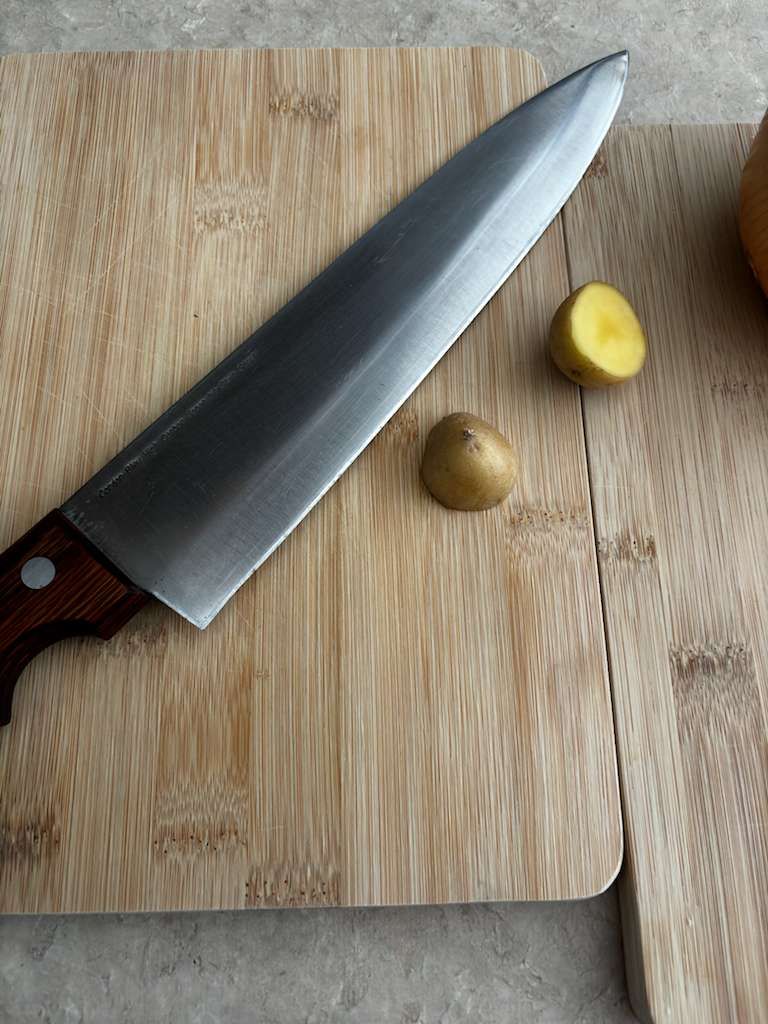 Chef's knife on a wooden cutting board with two small cut potatoes.