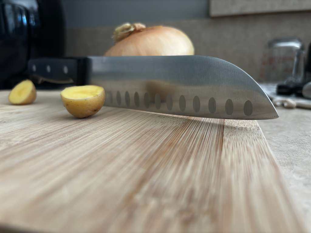 Knife on a wooden cutting board with two potato halves and an onion in the background.