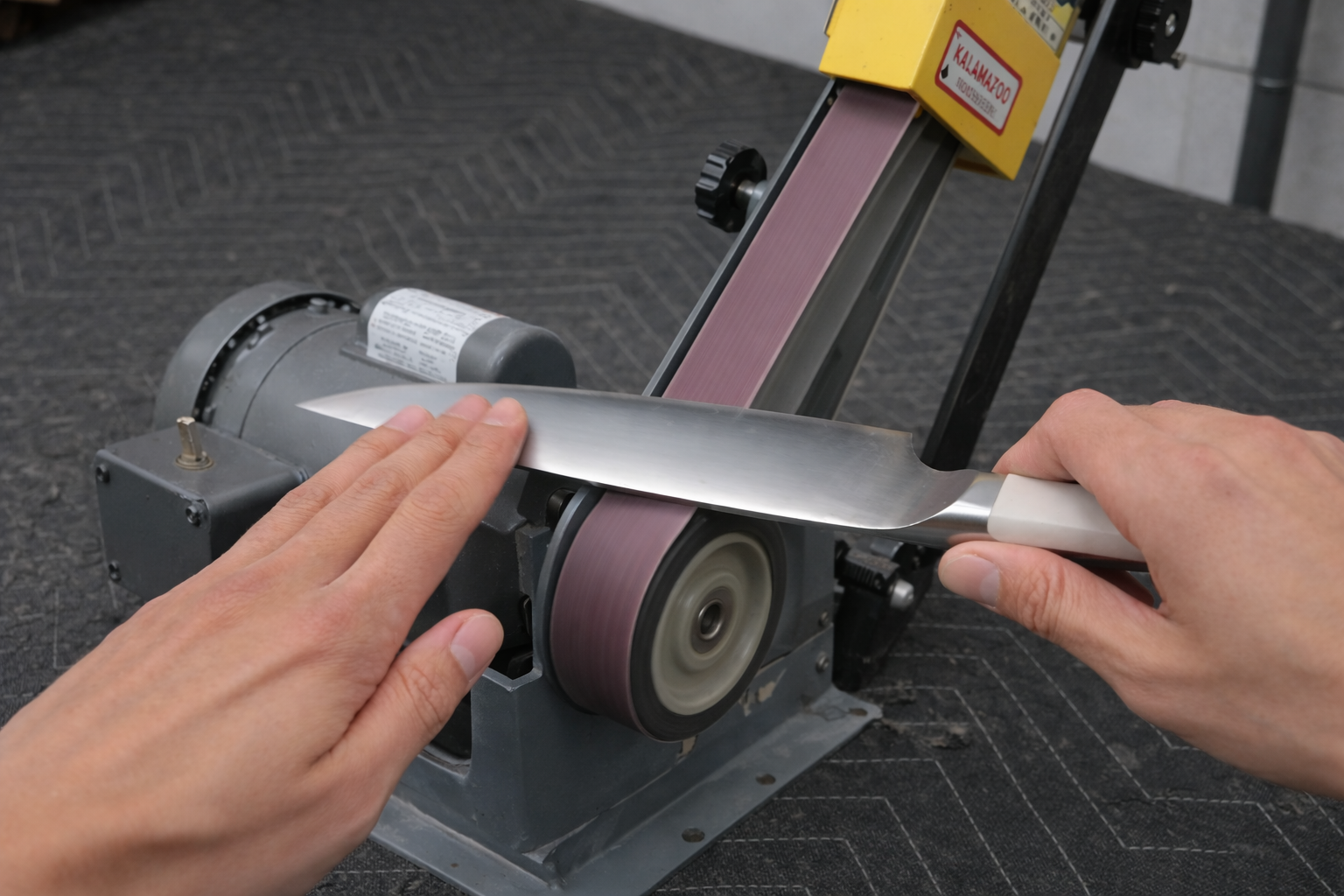 Person sharpening a knife blade on a belt grinder machine, hands guiding the blade, indoor setting.