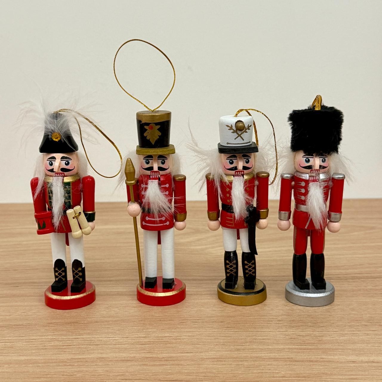 Cuatro adornos de cascanueces de madera en miniatura con uniformes rojos y blancos, sombreros negros y perchas de hilo dorado.