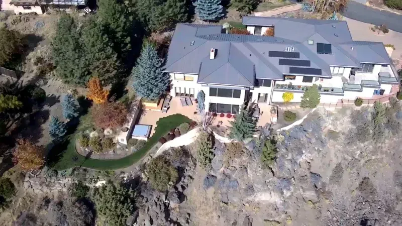 Large house with solar panels on a hillside, overlooking a valley with trees.