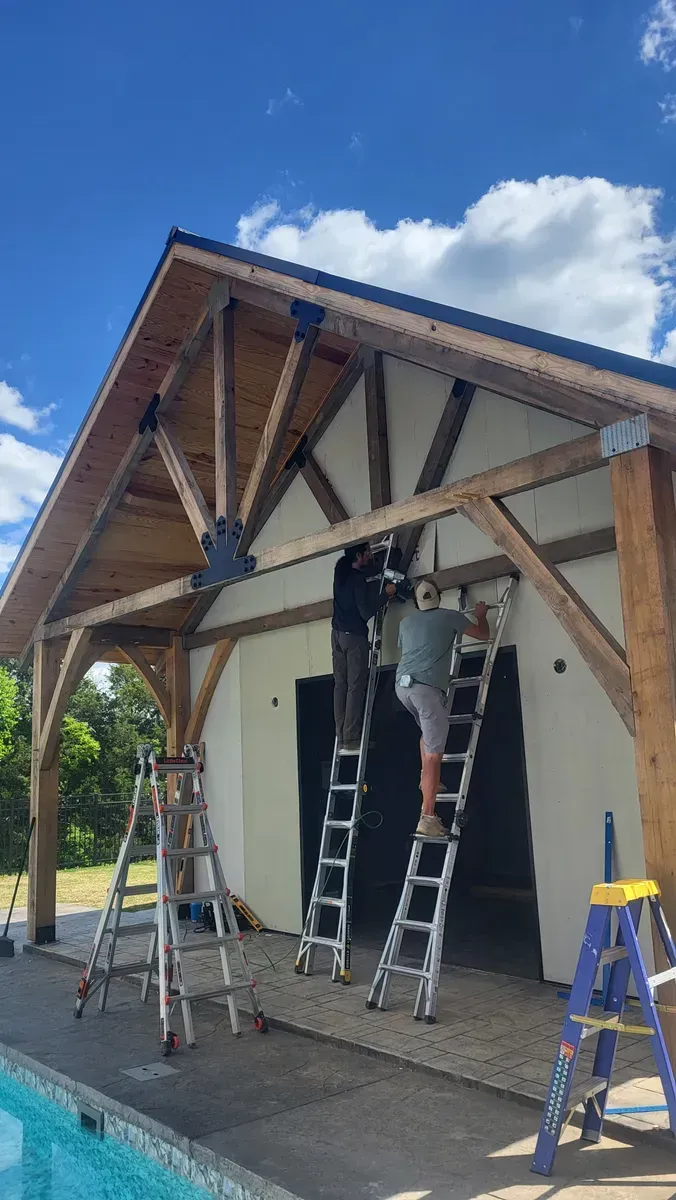 Two people on ladders working on a building with exposed wooden beams, near a pool, under a blue sky.