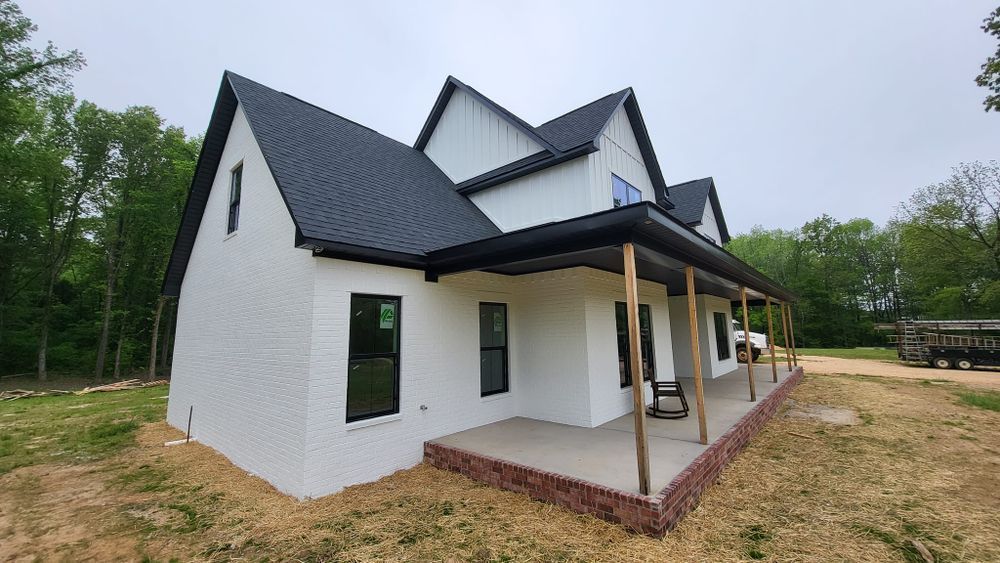 White farmhouse with a black roof and trim, and a porch, set in a wooded area.