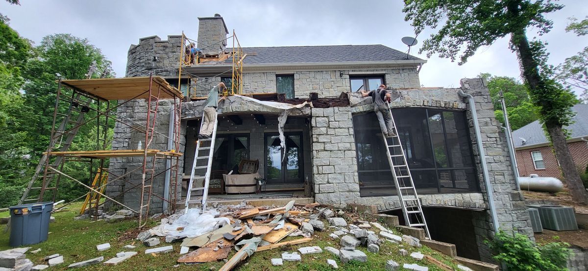House under construction, with scaffolding and ladders. Workers are visible, gray stone exterior. Cloudy sky.