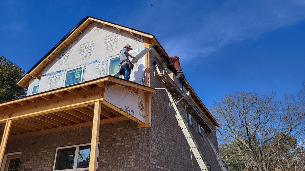 Construction workers on a ladder installing trim on a house under a clear blue sky.