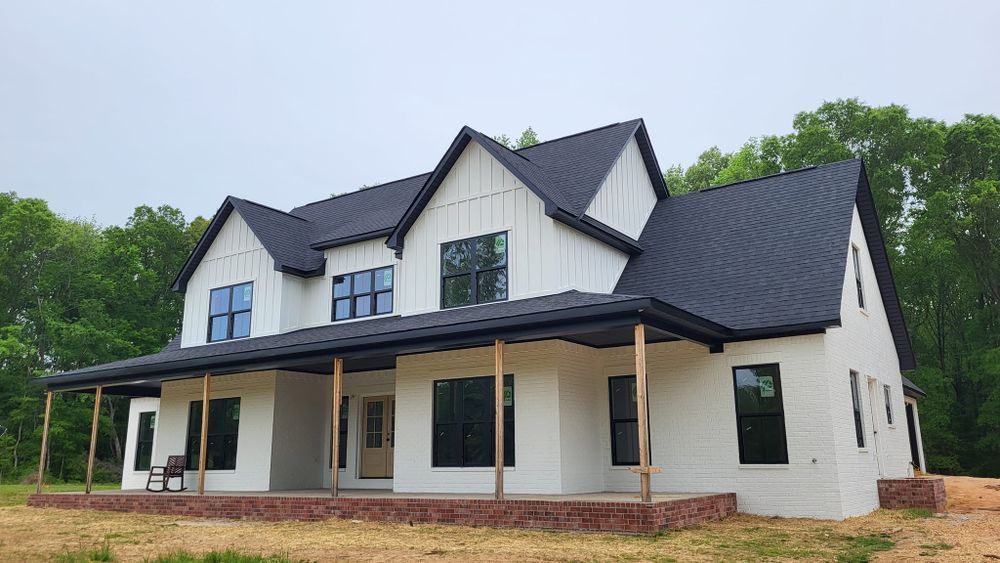 White farmhouse with black metal roof and trim, brick foundation, surrounded by trees.
