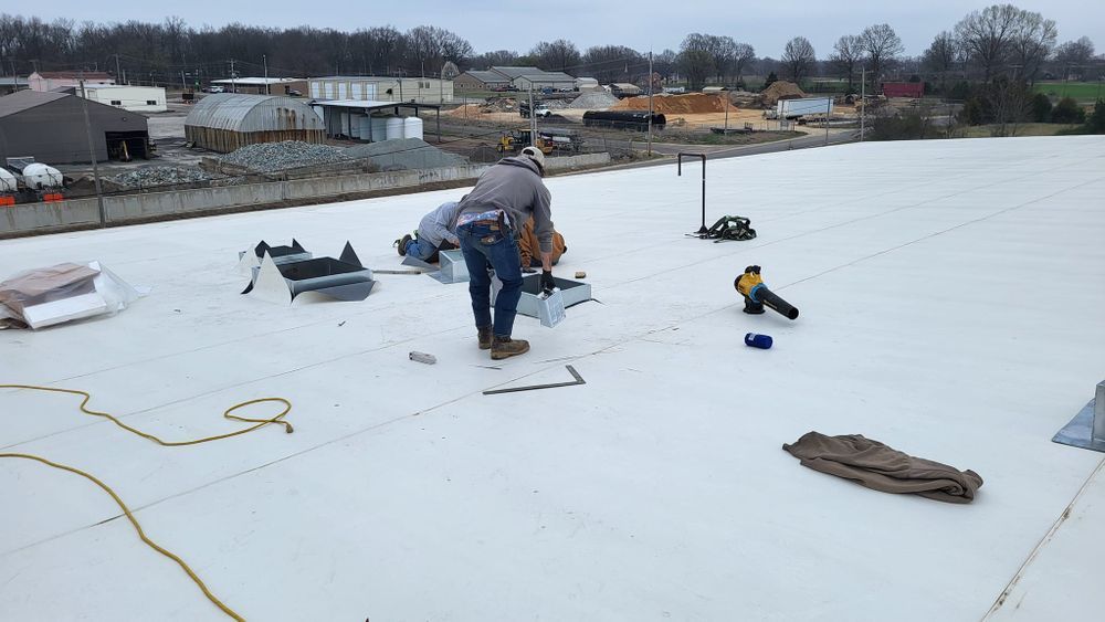 Roofer working on a white flat roof, using tools and materials. Buildings and cloudy sky in the background.