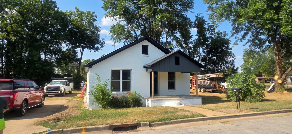 White house with dark trim and porch, parked vehicles in yard, trees overhead.