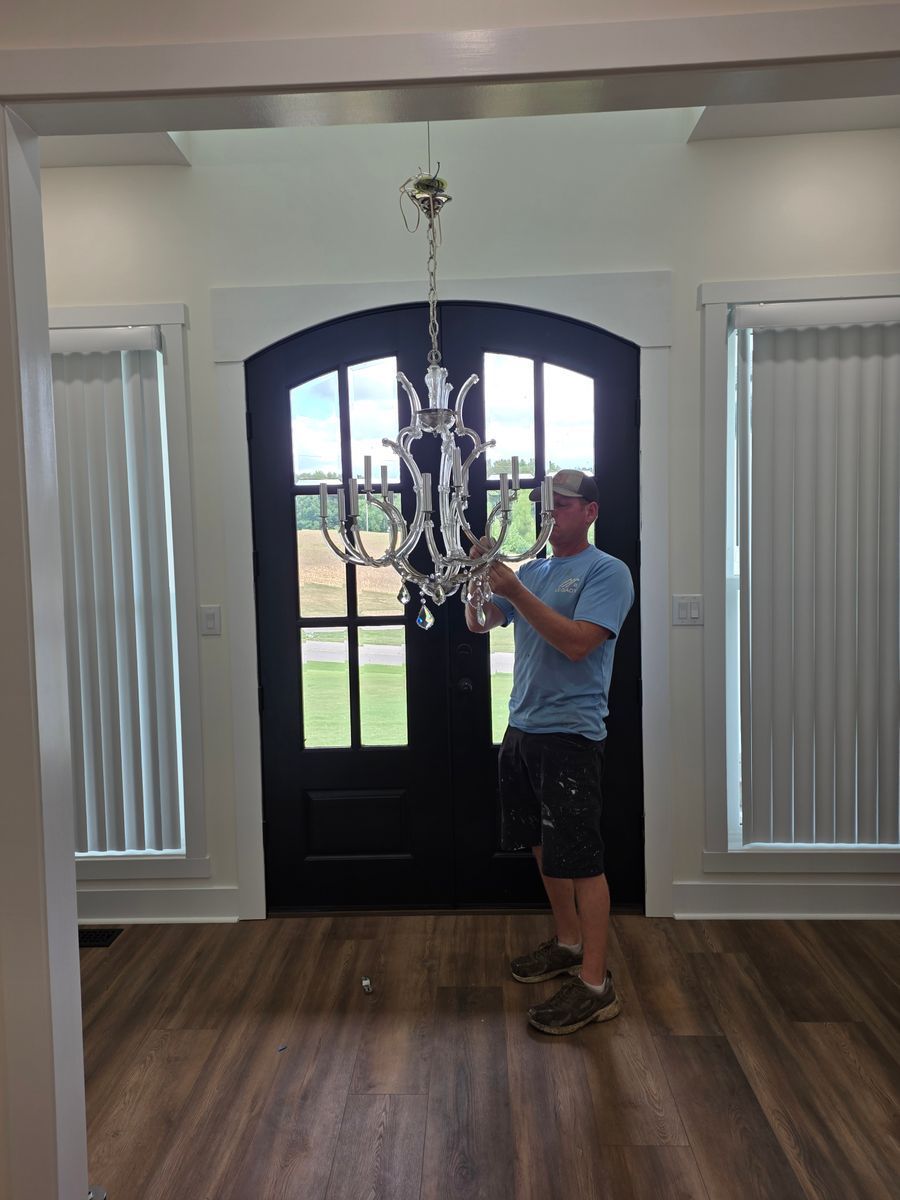 Man installing a crystal chandelier in a room with a black arched door, white trim, and wood flooring.