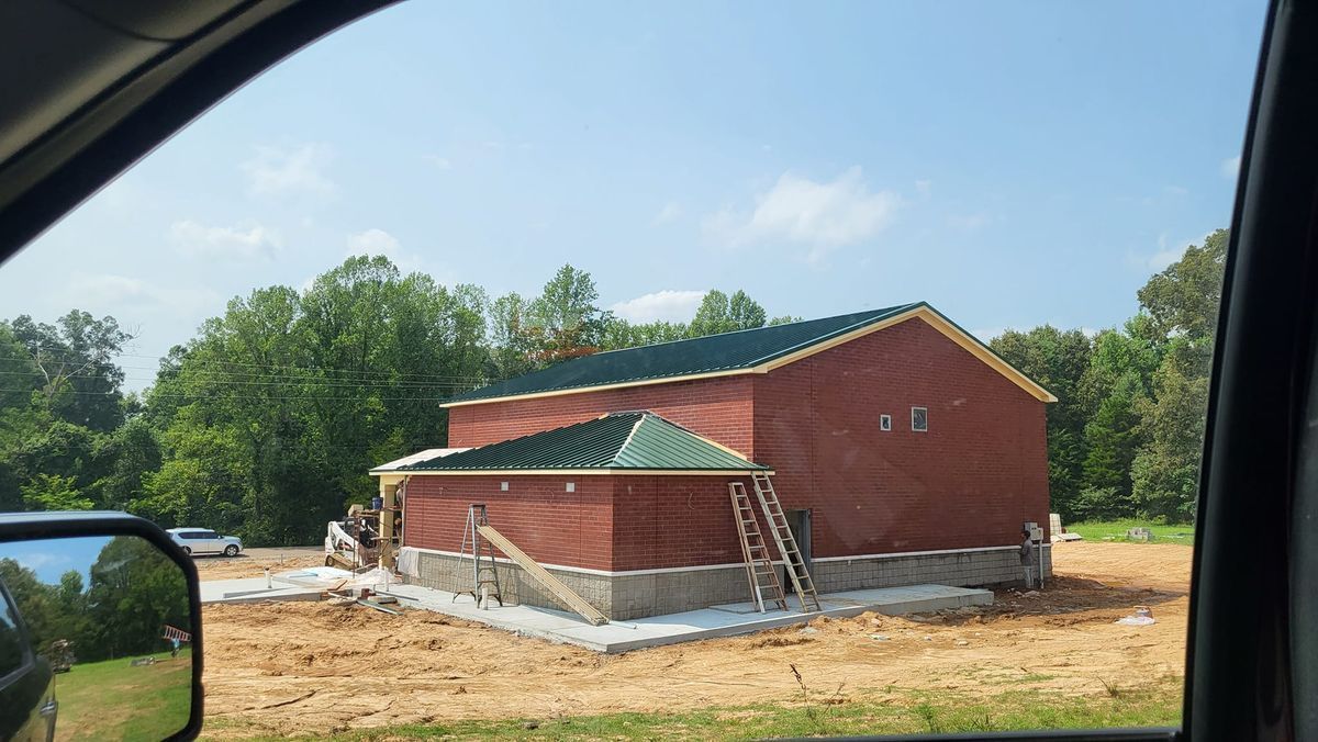Red brick building under construction with green metal roofs, sitting on a concrete foundation, trees in the background.