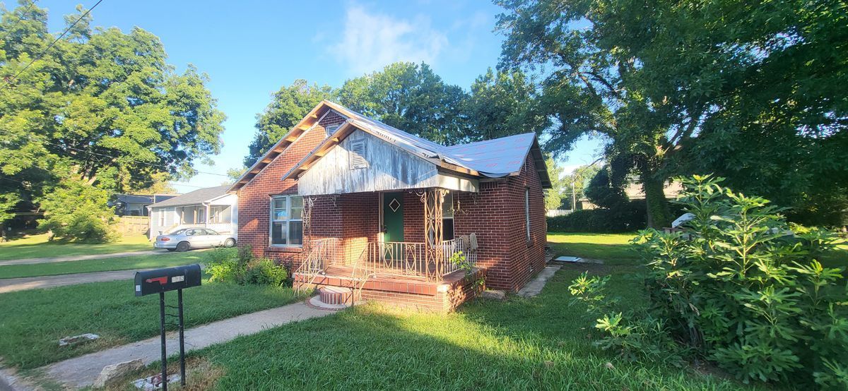 A weathered, wooden house with a metal roof sits on a grassy lot. A mailbox stands in front.