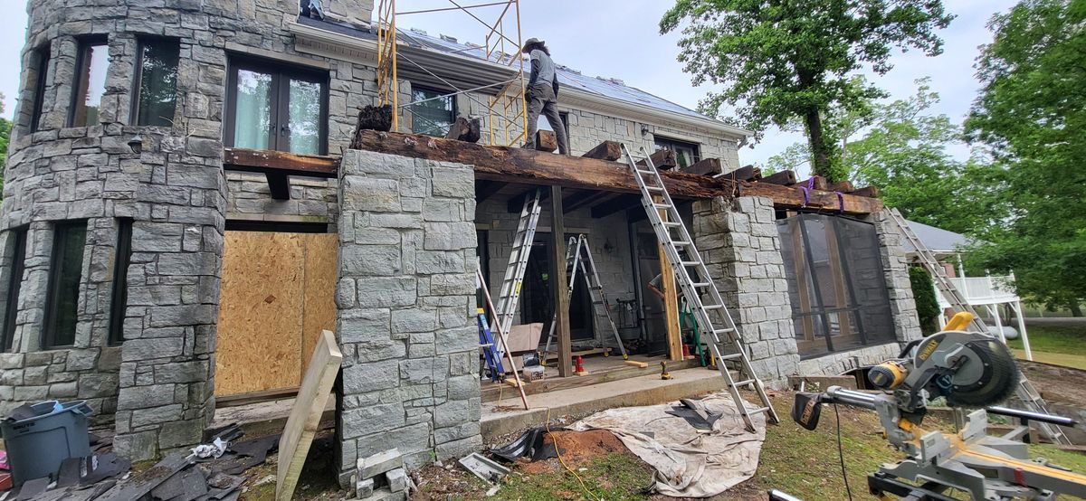 A stone house under construction, with a worker on the roof, ladders, and exposed beams.