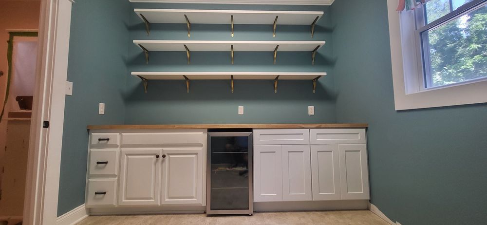 Room with teal walls, white cabinets, wooden countertop, and shelves. A wine fridge is centered below the shelves.